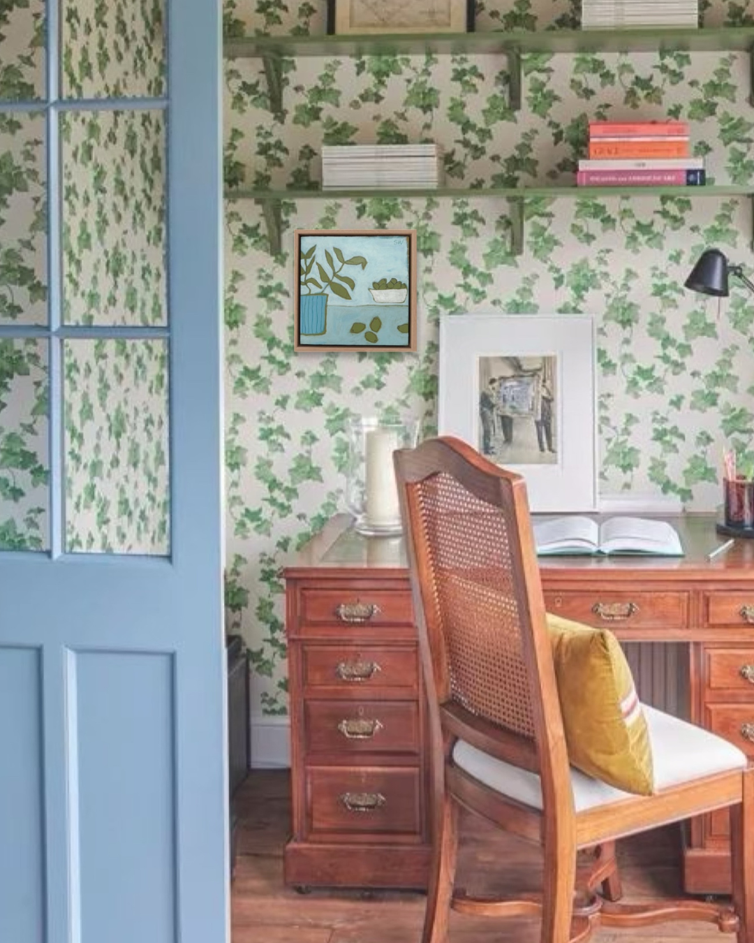 A cozy home office corner featuring green ivy patterned wallpaper, a dark wood writing desk, a woven-back cane chair with a mustard yellow pillow, and a light blue paneled door on the left.