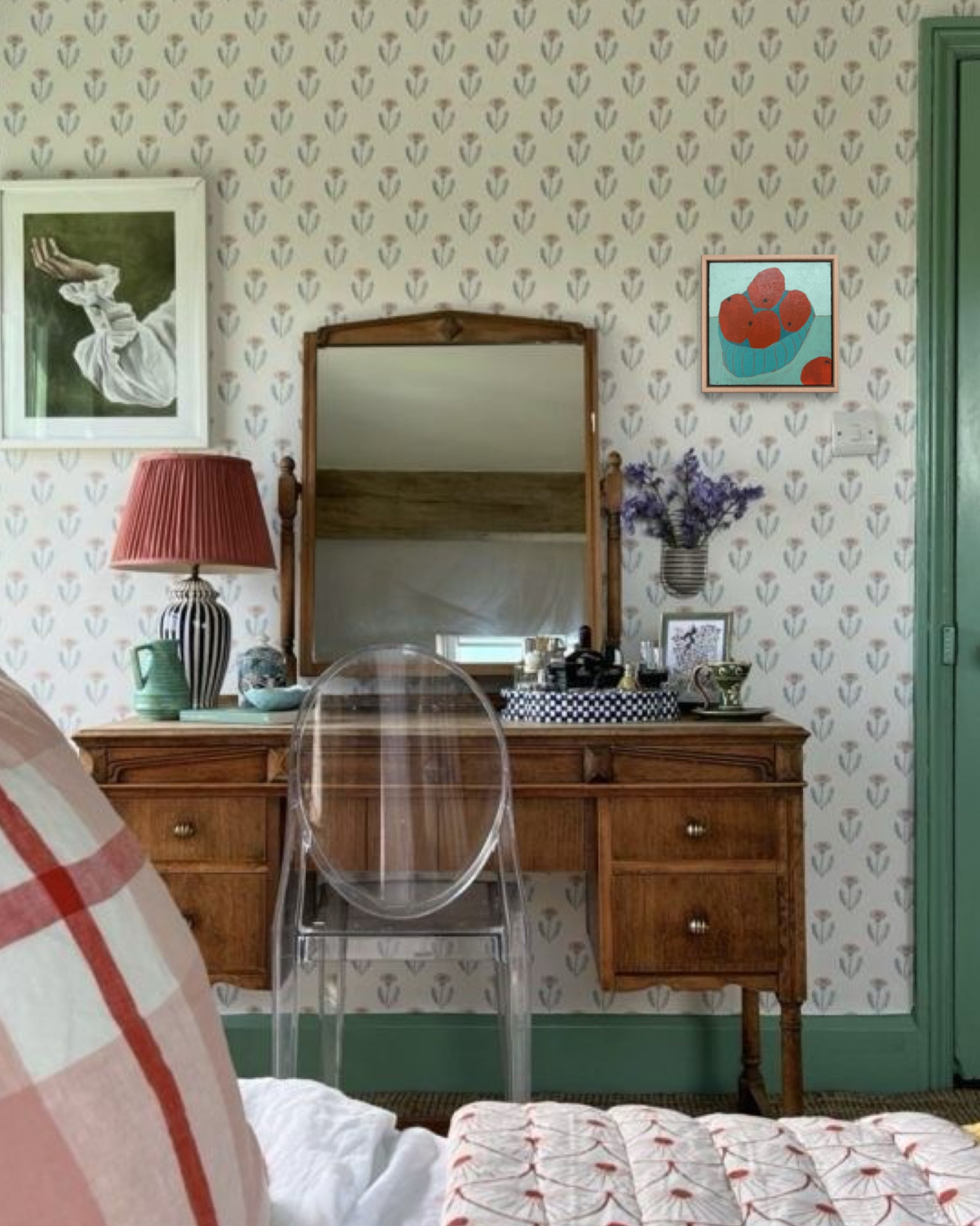 A vintage wooden writing desk with a large mirror, styled in a bedroom with patterned wallpaper, a red lamp, and a clear acrylic chair in the foreground.