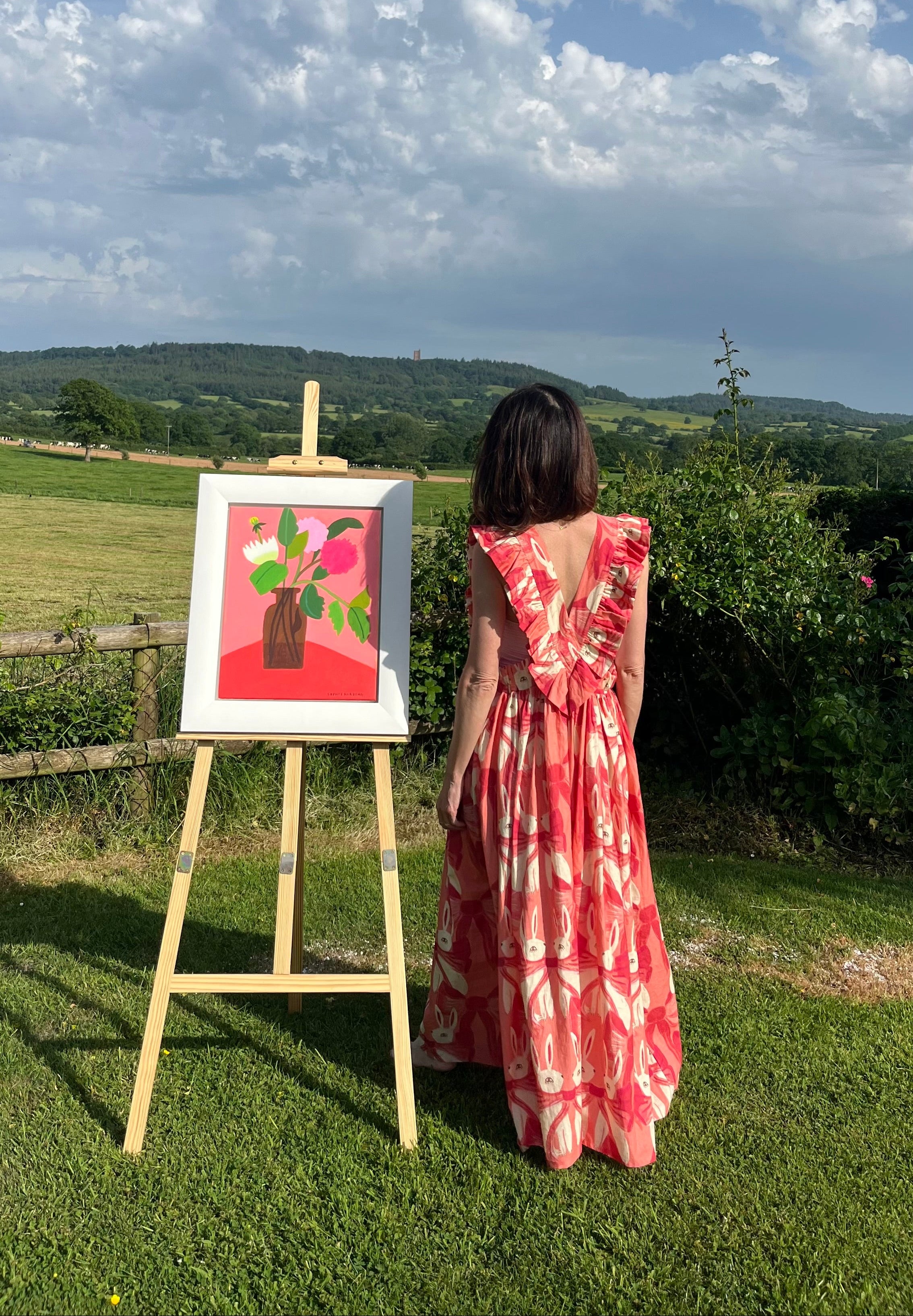 Woman in a red dress standing next to an easel with a painting outdoors