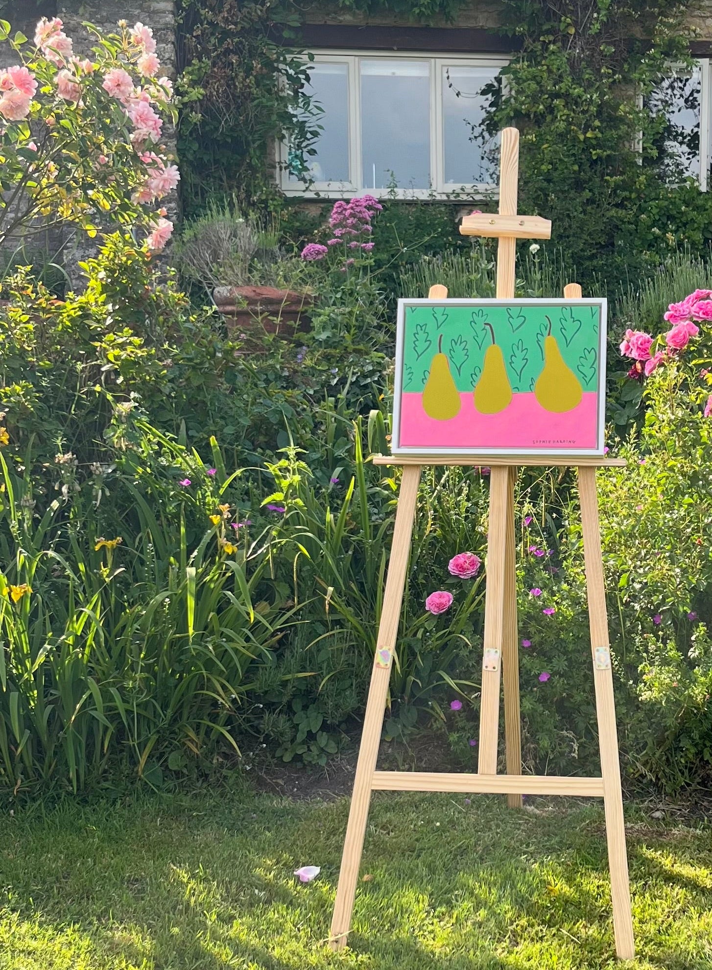 Painting of three yellow pears on a pink background displayed on an easel in a garden.