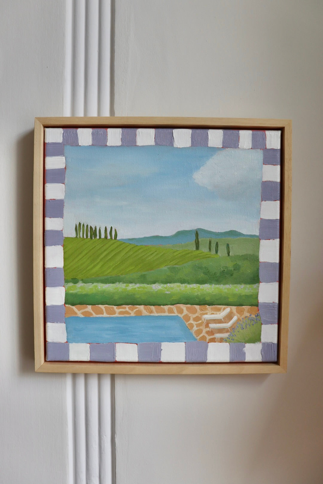 A framed landscape painting of a Tuscan vineyard and pool, featuring green rolling hills, cypress trees, a blue sky, and a decorative purple-and-white checkered border, mounted on a white paneled wall.
