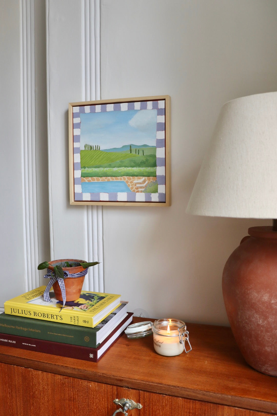 A framed landscape painting featuring green hills, a blue river, and a checkered border hangs on a light gray wall above a wooden cabinet. The cabinet holds a terracotta lamp base, a white lampshade, books, a small potted plant, and a lit candle.