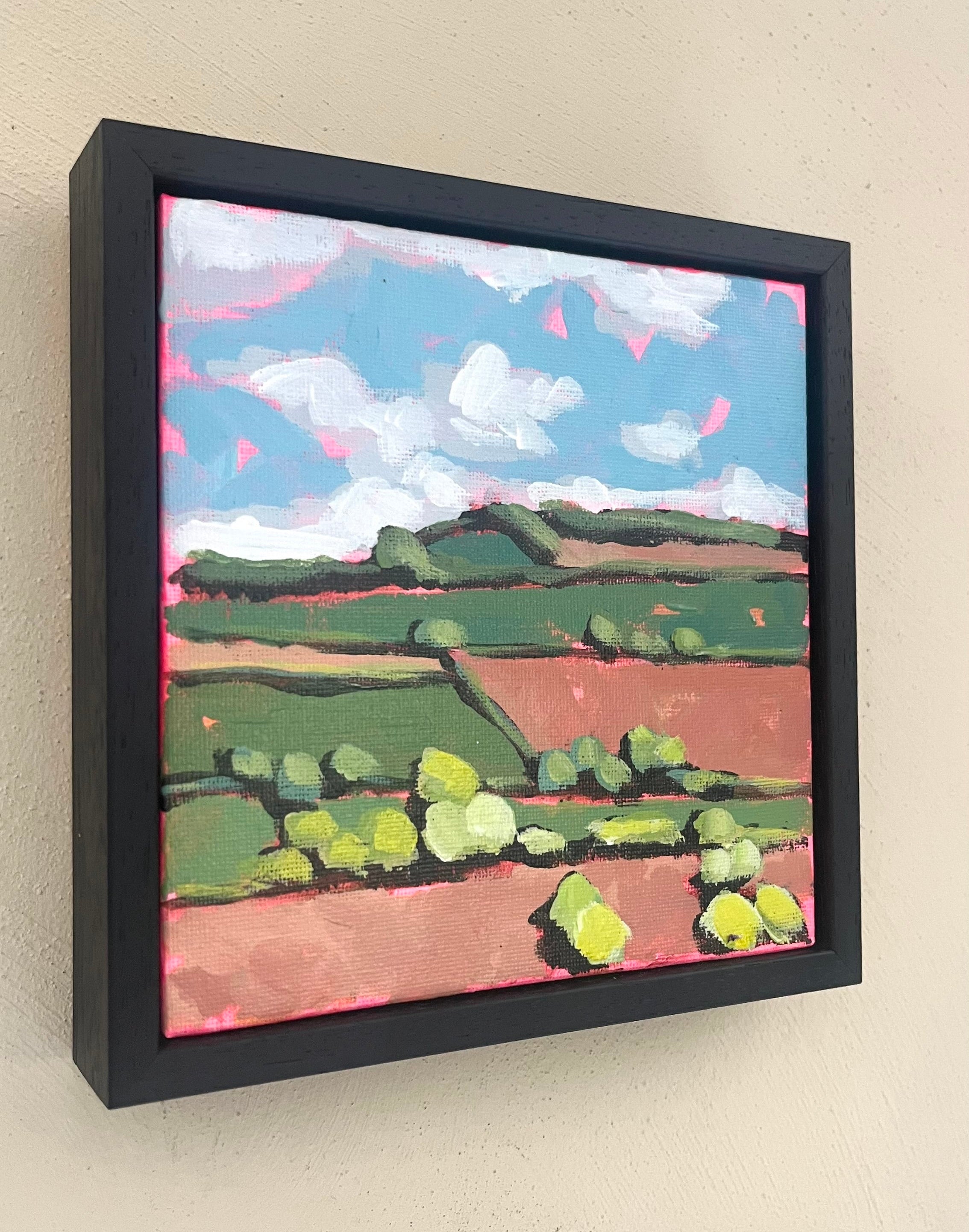 Framed painting of a landscape with green fields and blue sky on a beige wall.