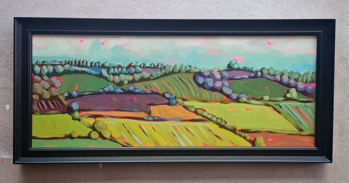 A framed landscape painting featuring colorful rolling hills, fields, and trees under a soft sky, enclosed in a black frame and mounted on a textured beige wall.