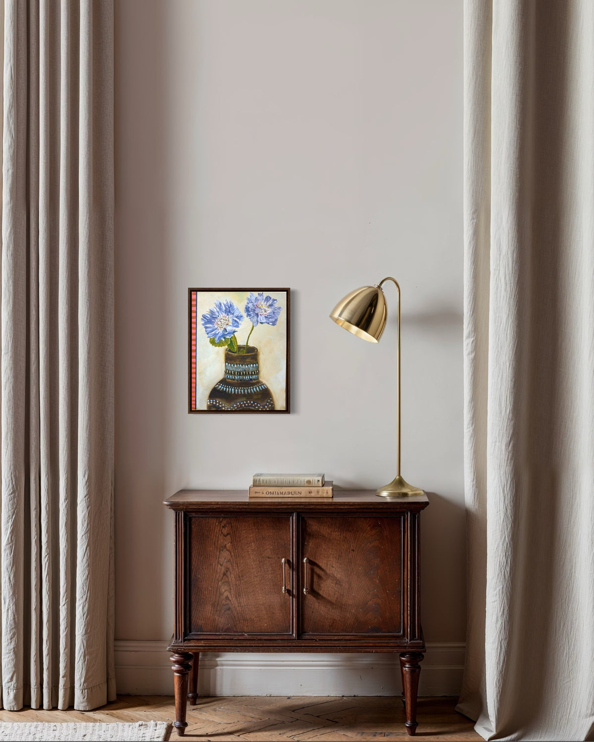 A styled interior scene with a dark wood cabinet against a beige wall, flanked by sheer curtains. On top of the cabinet sits a small framed floral painting, a stack of books, and a brass table lamp.
