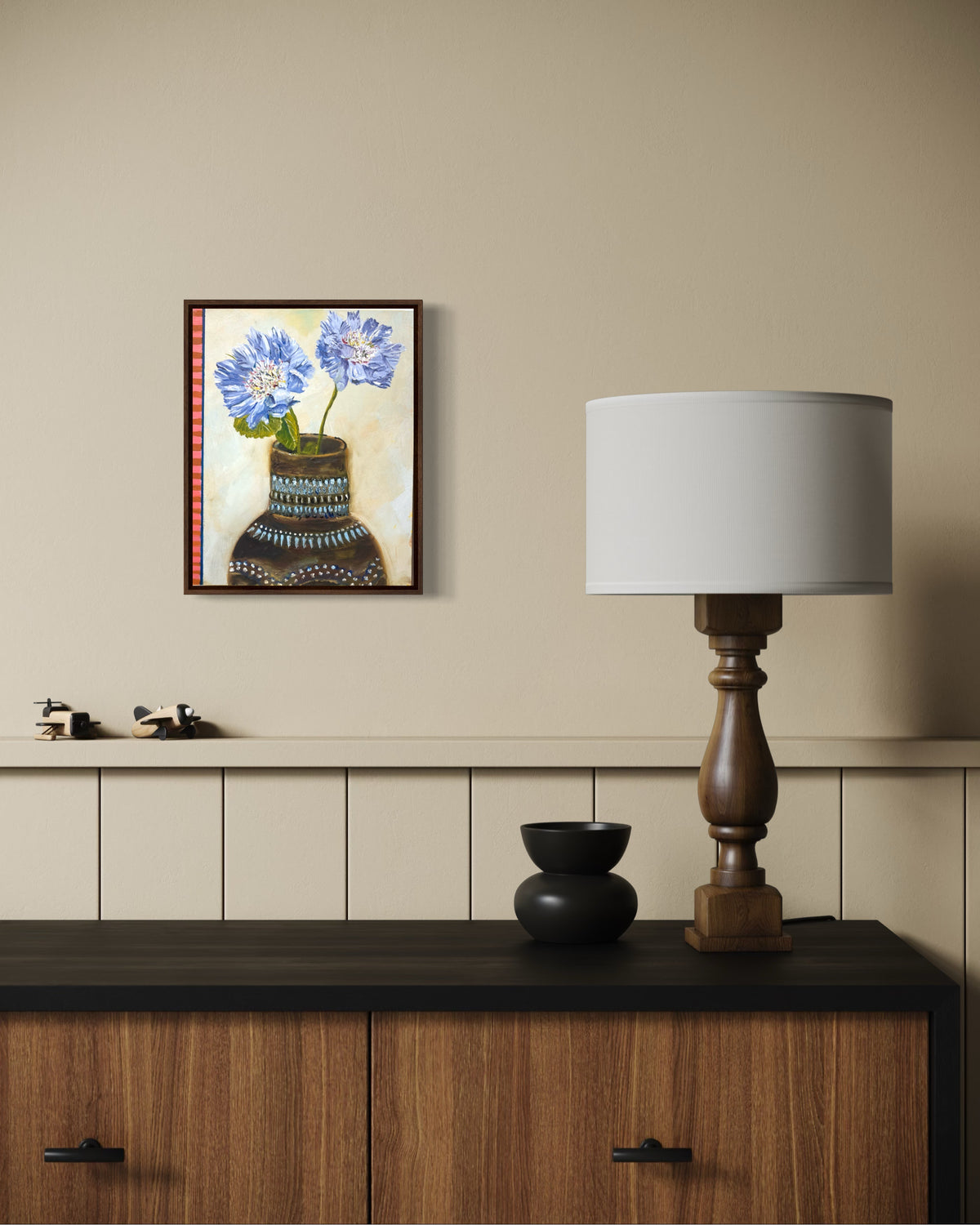 A framed art print depicting blue flowers in a patterned vase, displayed on a beige wall above a wooden cabinet. The scene is styled with a wooden table lamp and small decorative objects.